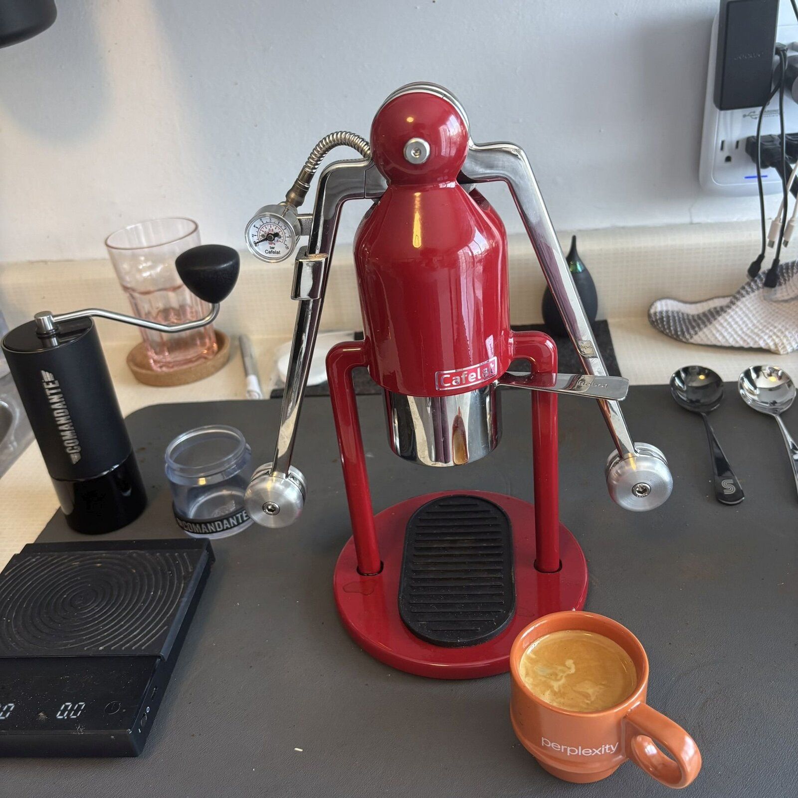 A red Cafelat Robot lever espresso machine next to a Comandante grinder and a scale, with a fresh espresso in an orange cup